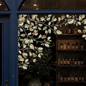 Könnte beinhalten: Ein Fensteraufkleber mit einer detaillierten Illustration von Eichenzweigen mit grünen Blättern und braunen Eicheln. Das Design ist auf dem Glas angebracht und zeigt ein natürliches botanisches Muster, das sich über das Fenster erstreckt.