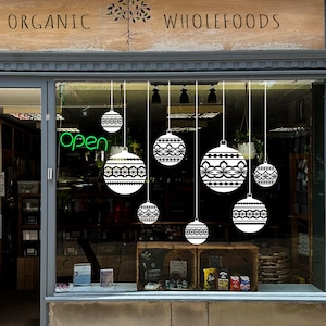 Könnte beinhalten: Ein Schaufenster zeigt hängende weiße und schwarze Zierkugeln. Das Schild darüber lautet "ORGANIC WHOLEFOODS". Ein neongrünes "Open"-Schild ist sichtbar. Das Fenster spiegelt das Innere des Geschäfts wider.