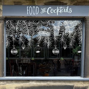Peut inclure: Une vitrine de magasin avec l'inscription "FOOD and Cocktails" en blanc. La fenêtre est ornée de dessins au trait blancs de branches de pin, de pommes de pin et de boules, créant un thème de vacances festif.