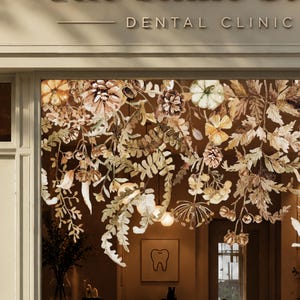 Op de afbeelding: Een etalage met een decoratieve herfststicker met pompoenen, dennenappels en bladeren in bruine, beige en crèmetinten. De tekst DENTAL CLINIC is boven het glas zichtbaar en binnen hangt een ingelijste illustratie van een tand.