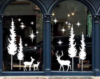 May include: A white vinyl window decal set featuring silhouettes of four evergreen trees, two deer, and one fawn, accented by various sizes of four-pointed stars. The design is applied to the glass panes of a dark-framed storefront window.