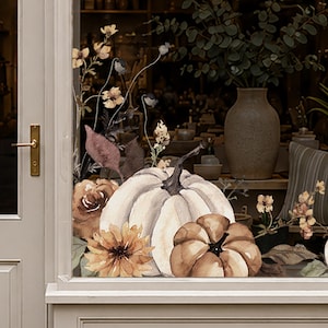 May include: A window decal featuring a large white pumpkin and a smaller brown pumpkin, surrounded by watercolour-style illustrations of autumn flowers, dried leaves, and thin stems, applied to the glass pane of a light-coloured door frame.