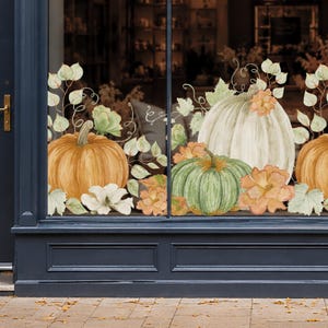 Peut inclure: Un autocollant de fenêtre présentant une illustration à l'aquarelle de citrouilles orange, vertes et blanches disposées avec des feuilles d'automne, des vignes et des fleurs. Le motif est appliqué sur un cadre de vitrine gris foncé.