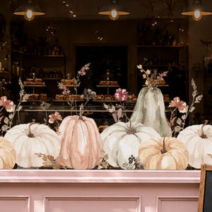 May include: A window decal featuring a watercolour illustration of various pumpkins in shades of cream, soft orange, and muted green, accented with delicate stems, dried wildflowers, and small floral elements arranged in a horizontal composition.