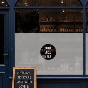Könnte beinhalten: Ein kreisförmiger schwarzer Fensteraufkleber mit dem weißen Text YOUR LOGO HERE, angebracht auf einer mattierten Glasscheibe. Unter dem Fenster zeigt ein hölzernes A-förmiges Kreidetafelschild den Text NATURAL SKINCARE MADE WITH LOVE &.