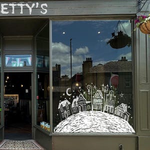 Könnte beinhalten: Außenansicht eines Schaufensters mit einer Kreidezeichnung einer Dorflandschaft. Das Geschäft heißt "Betty's". Die Zeichnung zeigt Häuser, einen Mond und Sterne. Das Fenster spiegelt den Himmel und die Gebäude wider.
