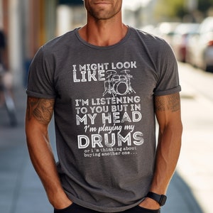 Pode incluir: Uma camiseta cinza com um gráfico branco de um kit de bateria e o texto "I might look like I'm listening to you but in my head I'm playing my drums or I'm thinking about buying another one..."