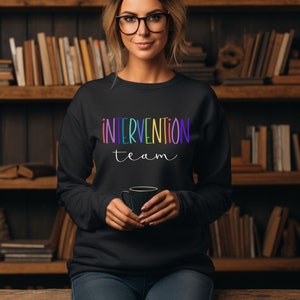 Puede incluir: Sudadera negra con las palabras "INTERVENTION team" en letras coloridas de arcoíris. La sudadera es de cuello redondo. El fondo es una estantería llena de libros.