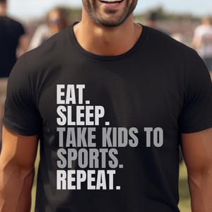 Op de afbeelding: Zwart T-shirt met ronde hals met de tekst "EAT. SLEEP. TAKE KIDS TO SPORTS. REPEAT." in witte en grijze letters. Het shirt wordt gedragen door een lachende persoon met een zonnebril.