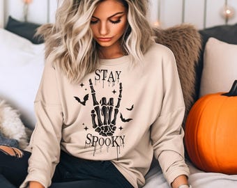 Może przedstawiać: Beżowa bluza z długim rękawem z czarnym napisem "STAY SPOOKY", przedstawiająca gest dłoni szkieletu i grafikę nietoperzy. Bluza ma luźny krój, idealna na Halloween.