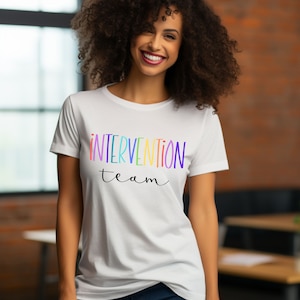 Puede incluir: Camiseta blanca con las palabras "INTERVENTION team" en letras coloridas de arcoíris. El texto está en un estilo manuscrito. La persona que lleva la camiseta está sonriendo.