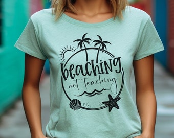 May include: A light green t-shirt with a black graphic design of palm trees, a sun, a seashell, a starfish, and the text "beaching not teaching".