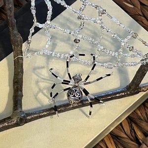 May include: A silver and black beaded spider ornament hanging from a white beaded web. The spider has a detailed design and is perched on a branch.