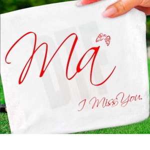 Puede incluir: Toalla cuadrada blanca con el texto rojo "Ma" y "I Miss You." Pequeñas alas de ángel rojas están encima del texto "Ma". La toalla está sujeta por una mano sobre un fondo verde.