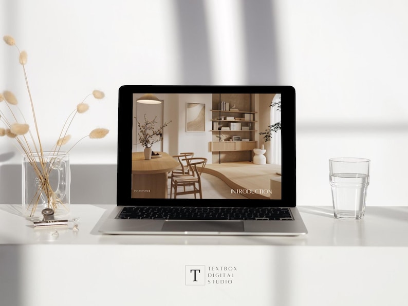 May include: A laptop displaying an interior design presentation with the word "INTRODUCTION" on the screen. The laptop is on a white surface, next to a glass of water and a vase with dried flowers. The Textbox Digital Studio logo is at the bottom.