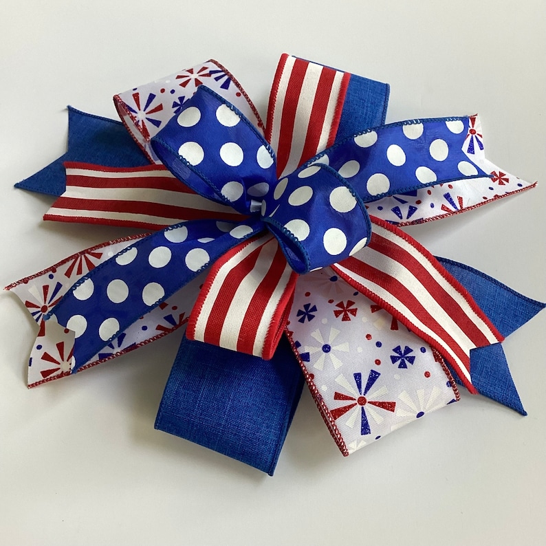 May include: A decorative bow featuring patriotic colors and patterns. The bow has blue, red, and white ribbons with polka dots, stripes, and starburst designs. The bow is suitable for wreaths, gifts, or holiday decorations.