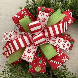 May include: A festive Christmas bow with red and white striped ribbon, green ribbon, and red ribbon with candy cane and peppermint patterns. The bow is set against a backdrop of green foliage.