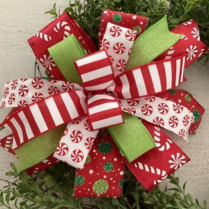 May include: A festive Christmas bow with red and white striped ribbon, peppermint candy print ribbon, and green accents. The bow is decorated with glitter and is set against a backdrop of greenery.