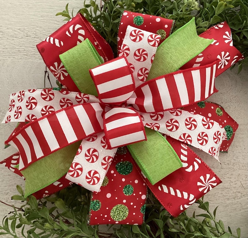 May include: A festive Christmas bow with red and white striped ribbon, peppermint candy print ribbon, and green accents. The bow is decorated with glitter and is set against a backdrop of greenery.