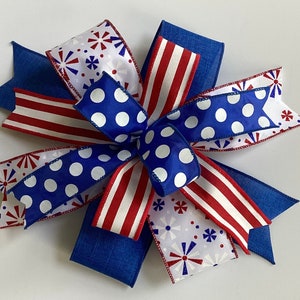 May include: A decorative bow featuring patriotic colors and patterns. The bow has blue, red, and white ribbons with polka dots, stripes, and starburst designs. The bow is made of fabric and is suitable for various craft projects.