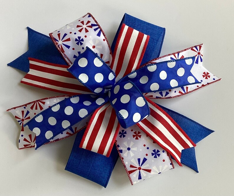 May include: A decorative bow featuring patriotic colors and patterns. The bow has blue, red, and white ribbons with polka dots, stripes, and starburst designs. The bow is made of fabric and is suitable for various craft projects.