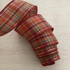 May include: A roll of wired ribbon featuring a plaid pattern in shades of burnt orange, deep red, forest green, and dark navy blue. The fabric is accented with thin, metallic gold horizontal and vertical lines that create a grid across the surface.