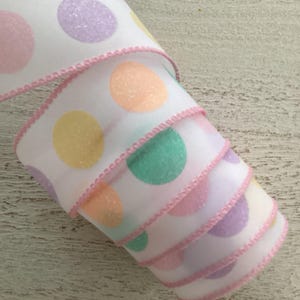 Peut inclure: Ruban blanc à pois pastel roses, violets, jaunes et verts. Le ruban a un bord cousu rose. Le ruban est enroulé, montrant plusieurs couches du motif.