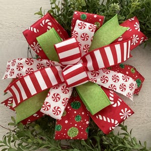 May include: A festive Christmas bow featuring red and white striped ribbon, green ribbon, and ribbon with candy cane and peppermint designs. The bow is accented with glitter and set against a backdrop of greenery.