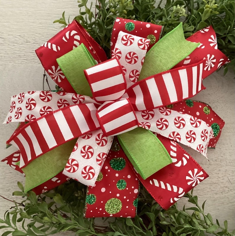 May include: A festive Christmas bow featuring red and white striped ribbon, green ribbon, and ribbon with candy cane and peppermint designs. The bow is accented with glitter and set against a backdrop of greenery.