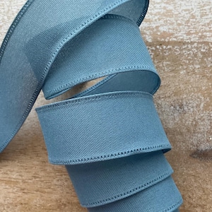 May include: A roll of solid blue ribbon with a stitched edge. The ribbon is a muted teal color and appears to be made of a woven fabric. The ribbon is curled in a spiral, showing the texture and detail of the material.