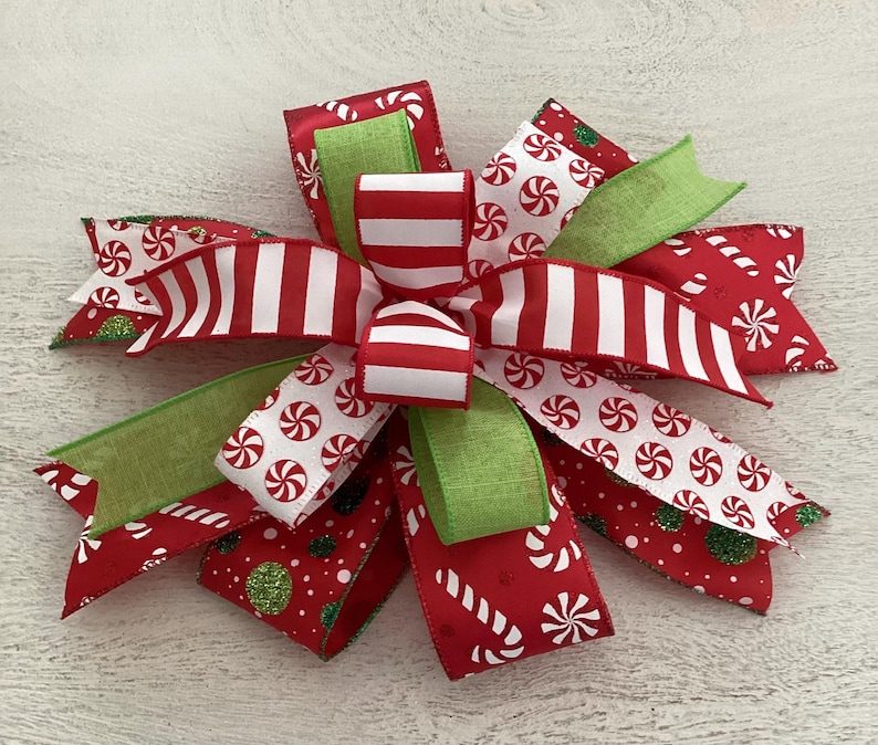 May include: A festive Christmas bow featuring red and white striped ribbon, green ribbon, and red ribbon with candy cane and peppermint patterns. The bow is a decorative accent for holiday decor.