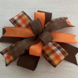 May include: A decorative bow featuring a mix of brown, orange, and plaid ribbons. The bow has a layered design with various ribbon textures and patterns, perfect for fall-themed crafts and decorations.