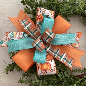 May include: A decorative bow featuring orange, teal, and plaid ribbons. The bow has a multi-layered design with pumpkin and berry patterns. The bow is set against a backdrop of green foliage, perfect for fall decor.