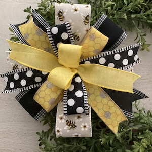 May include: A decorative bow featuring multiple ribbon layers in yellow, black with white polka dots, and a beige pattern with bees and white flowers. The centre is a solid yellow ribbon, with additional yellow honeycomb patterned ribbons.