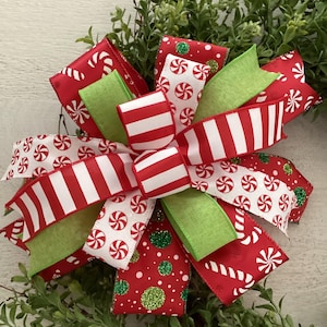 May include: A festive Christmas bow with red and white striped ribbon, peppermint candy print ribbon, and green ribbon accents. The bow is set against a backdrop of green foliage, creating a cheerful holiday decoration.