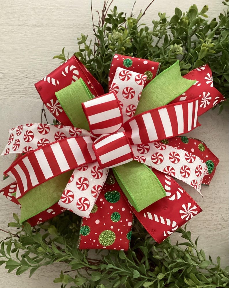 May include: A festive Christmas bow with red and white striped ribbon, peppermint candy print ribbon, and green ribbon accents. The bow is set against a backdrop of green foliage, creating a cheerful holiday decoration.