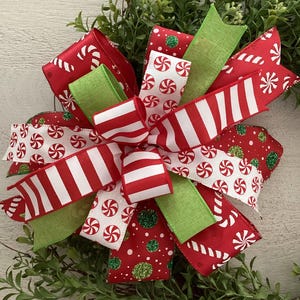 May include: A festive Christmas bow featuring red and white striped ribbons, candy cane patterned ribbons, and ribbons with white peppermint candies on a red background. Green ribbons and glitter accents add to the holiday theme.