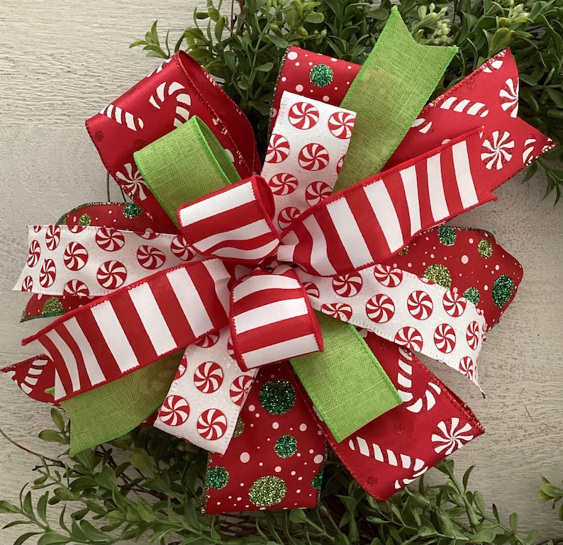 May include: A festive Christmas bow featuring red and white striped ribbons, candy cane patterned ribbons, and ribbons with white peppermint candies on a red background. Green ribbons and glitter accents add to the holiday theme.