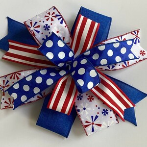 May include: A decorative bow featuring patriotic colors and patterns. The bow has blue, white, and red ribbons. Some ribbons have white polka dots on a blue background, while others have red and white stripes or a firework design.