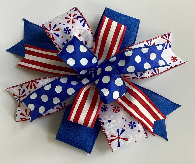 May include: A decorative bow featuring patriotic colors and patterns. The bow has blue, white, and red ribbons. Some ribbons have white polka dots on a blue background, while others have red and white stripes or a firework design.