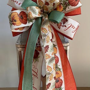 May include: A decorative bow made of wired ribbon featuring a pattern of orange pumpkins and autumn leaves on a cream background, accented with solid burnt orange and sage green ribbons. The text "Thankful" is printed in a cursive script on the ribbon.