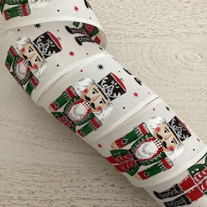 May include: White ribbon with a repeating pattern of toy nutcracker soldiers in green, red, and black uniforms. The ribbon also has small red and black star accents. The ribbon is rolled up, showing the pattern.