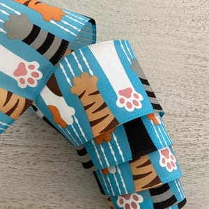 May include: Decorative ribbon with a repeating pattern of cat paws and tiger stripes. The ribbon has a light blue background with white vertical stripes. The cat paws are pink, and the tiger stripes are brown and black.