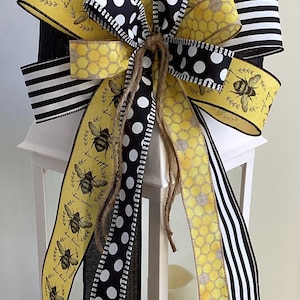 May include: A white lantern with a yellow, black, and white bee-themed bow. The bow is made of four different ribbons: one with a honeycomb pattern, one with black and white polka dots, one with black and white stripes, and one with bees and the text "bee happy, bee sweet, bee loved".
