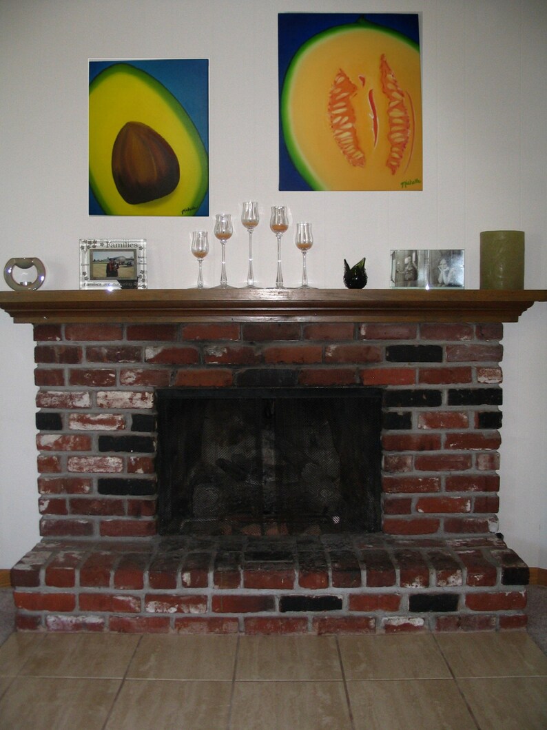 May include: A brick fireplace with a black metal fire screen. Above the fireplace are two framed paintings, one of a cantaloupe and one of an avocado. There are also four clear glass candle holders on the mantel.