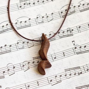 Puede incluir: Un colgante de madera marrón en forma de nota musical en un cordón de cuero marrón. El colgante está colgando sobre una hoja de música.