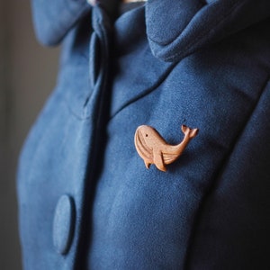 Puede incluir: Un broche de madera con forma de ballena prendido en una chaqueta azul oscuro. La ballena está tallada en un estilo simple y minimalista.