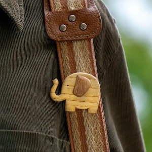 Puede incluir: Un broche de madera con forma de elefante, marrón claro con una oreja más oscura, está sujeto a una correa a rayas con un acento de cuero marrón. La correa es de color beige y marrón. El broche está sujeto a una camisa de pana verde.