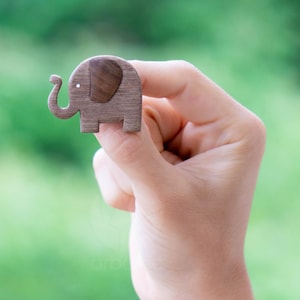 Puede incluir: Una pequeña figura de elefante de madera con una oreja marrón oscuro y un punto blanco para el ojo. El elefante es de color marrón claro y tiene una trompa curvada. La figura se sostiene en una mano sobre un fondo verde borroso.