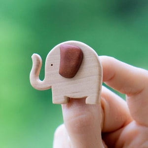 Puede incluir: Una pequeña figura de elefante de madera con un cuerpo marrón claro y una oreja marrón oscuro. El elefante tiene una trompa curvada. El fondo es verde borroso.